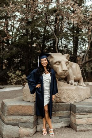 A graduate in academic regalia poses next to a stone lion statue in an outdoor setting with trees in the background.