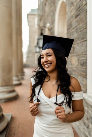 Graduate in white dress and graduation cap smiles joyfully while posing against stone wall background during photo session.