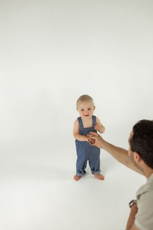 Small child in blue denim overalls stands barefoot on white background while reaching for adult's extended hand.