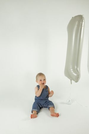 Baby in denim overalls crawls and plays on white studio backdrop with silver balloon.