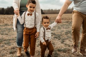 Two small children in matching suspender outfits walk hand in hand with adults in a field.