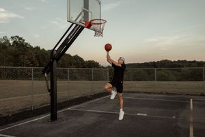 Basketball player jumps high for a layup on an outdoor court at sunset with trees in the background.