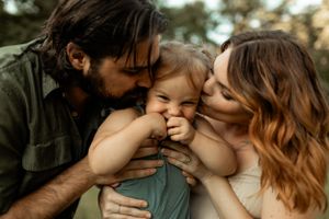 Tender family moment captured outdoors during golden hour with parents kissing child's cheeks.