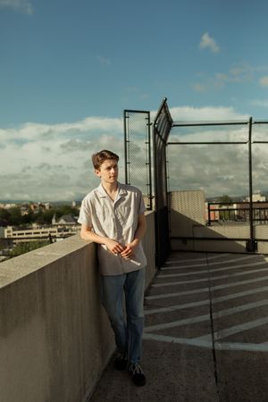 Person in white shirt and jeans poses on a rooftop parking structure against an urban backdrop at sunset.