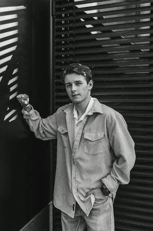 Black and white photo showing a person in a light jacket posing expressively against a slatted background.