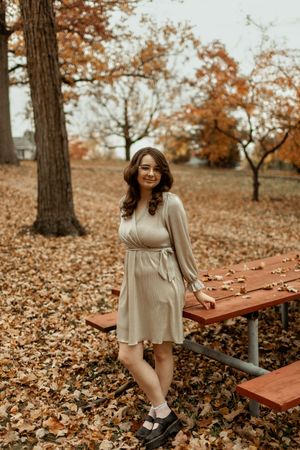 A person sits on a wooden picnic table surrounded by fallen autumn leaves in a park setting.