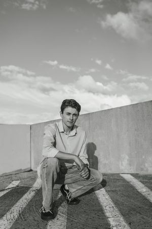 Black and white photograph of someone sitting on a rooftop against a wall with cloudy sky in the background.