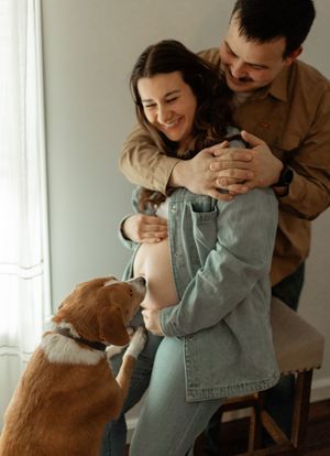 A couple shares a tender moment with their dog by a bright window as they embrace in casual attire.