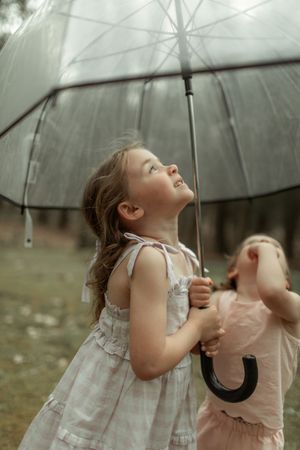 Young figure in a white dress twirls with an umbrella outside while looking up at a cloudy sky with joy and wonder.