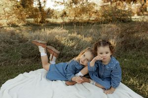 Two children in denim outfits lay together on a white blanket in a grassy outdoor setting at golden hour.