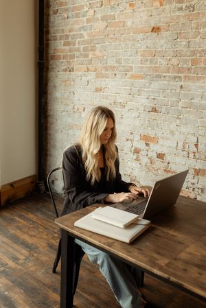 Person in black blazer working at wooden desk with laptop and book against exposed brick wall.