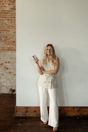 Person in white outfit standing against brick wall while looking at mobile phone.