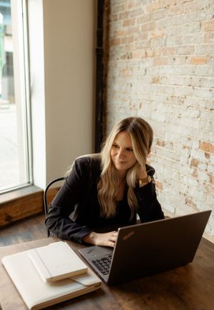 Person working on laptop at wooden desk near exposed brick wall in natural lighting from window.