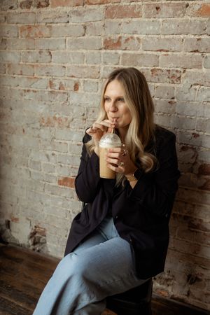 A person in a black sweater drinks from a white mug while sitting against a rustic brick wall.