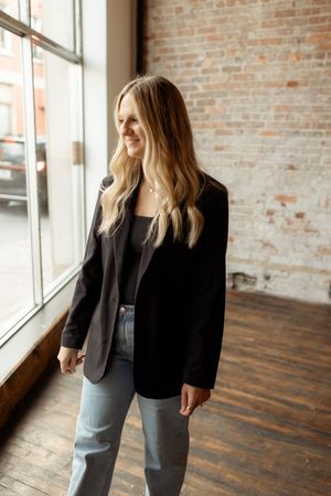 A professional wearing a black blazer and light jeans stands near a window with exposed brick walls.
