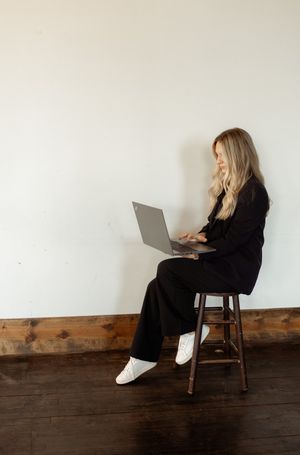 A minimalist workspace setup with someone working on a laptop while seated on a wooden stool.