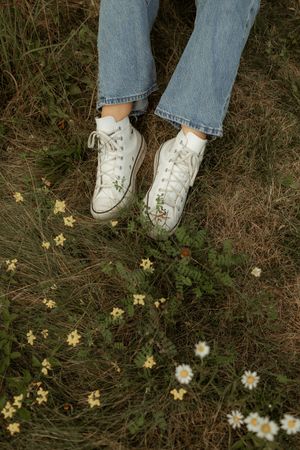 A person wearing blue jeans and white Converse sneakers stands among small yellow wildflowers in grass.