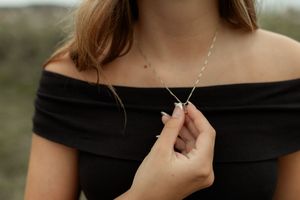 Close-up of hands holding delicate silver necklace pendant against black off-shoulder top in outdoor setting.