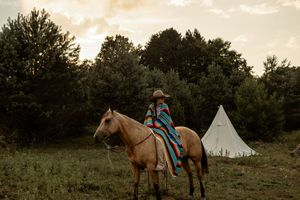 A rustic countryside scene with a grazing horse and tipi tent silhouetted against a warm sunset sky.