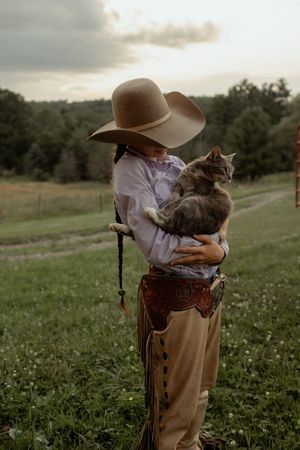 A person in a sun hat and white shirt holds a gray cat while standing in a wildflower meadow at sunset.