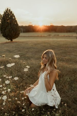 A person in a flowing white dress sits in a golden field of wildflowers at sunset.