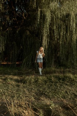 A person stands beneath a dramatic weeping willow tree in summer wearing denim shorts and white boots.