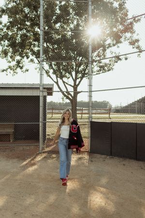 Person walking through a sunlit baseball field with chain link fences and trees casting long shadows at sunset.