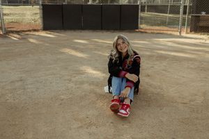 A person in a denim jacket and backpack sits on a concrete curb in an outdoor setting.