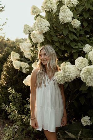 A person in a white flowy top poses against blooming white hydrangea bushes during golden hour.