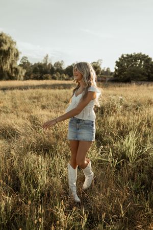 A person wearing denim shorts and white boots walks through a golden field at sunset.