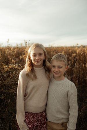 Two young people in beige sweaters pose together against an autumn field at sunset.