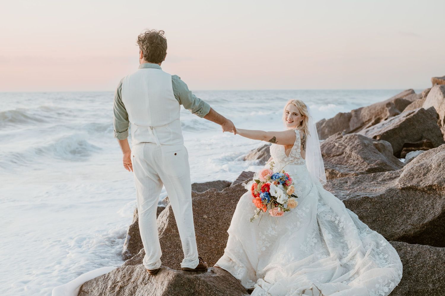 A wedding couple stands on rocky coastal outcrop at sunset holding hands with crashing waves in background.