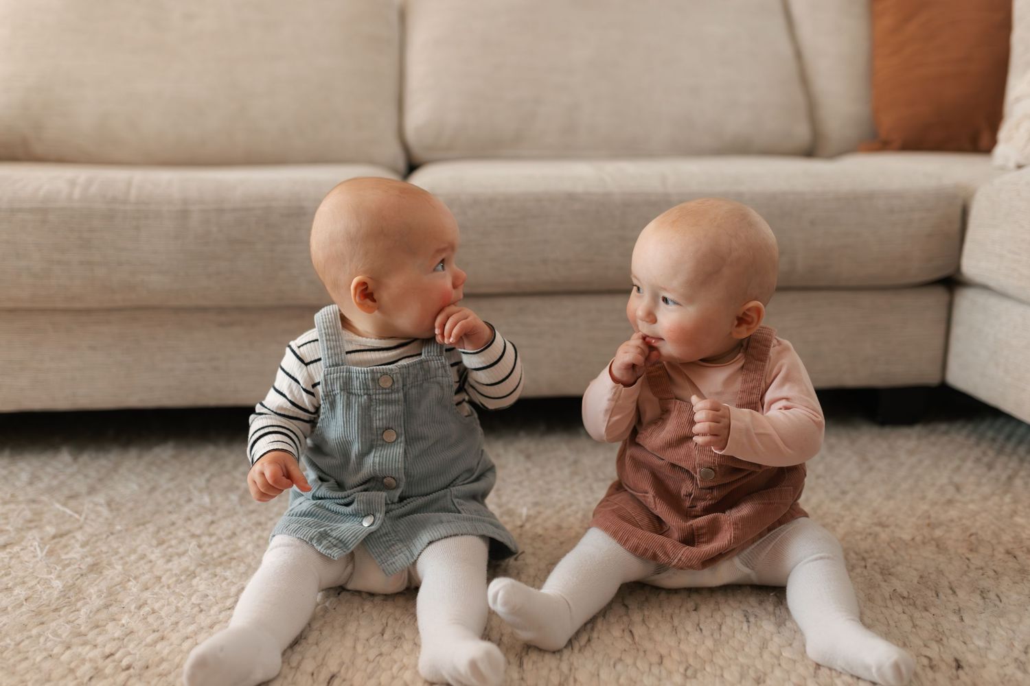Two adorable bald babies in overalls sitting on carpet facing each other near beige couch.