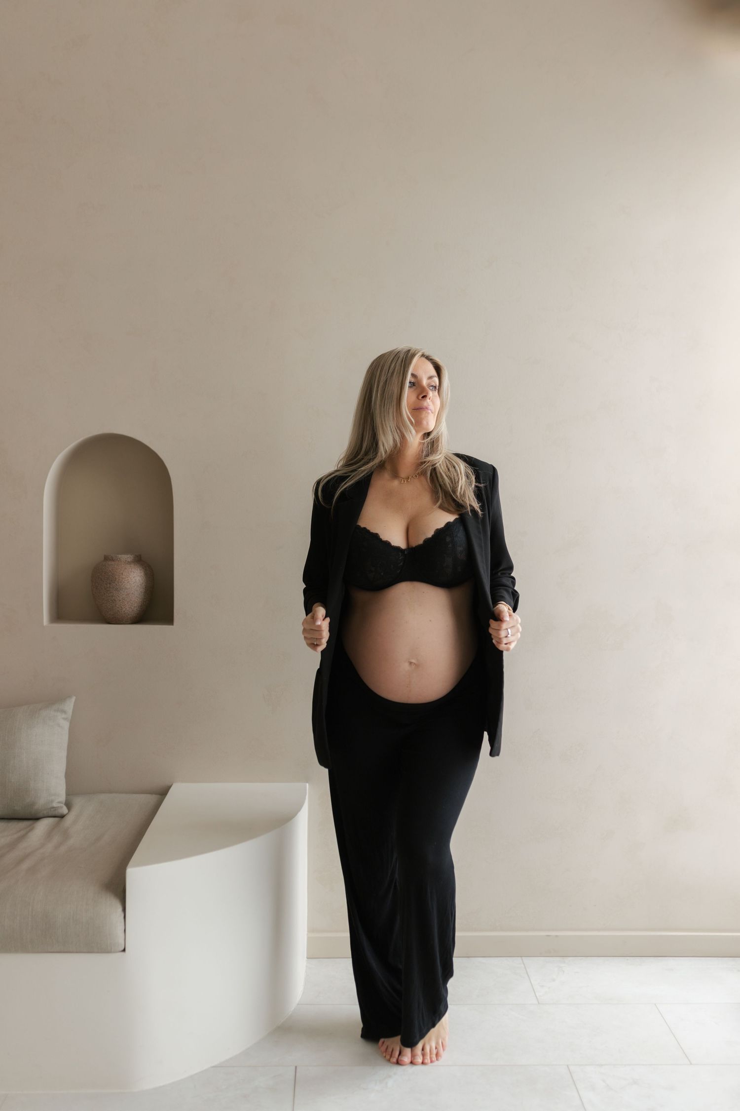 Pregnant woman in black blazer and lace bralette showcasing baby bump in minimalist neutral interior.