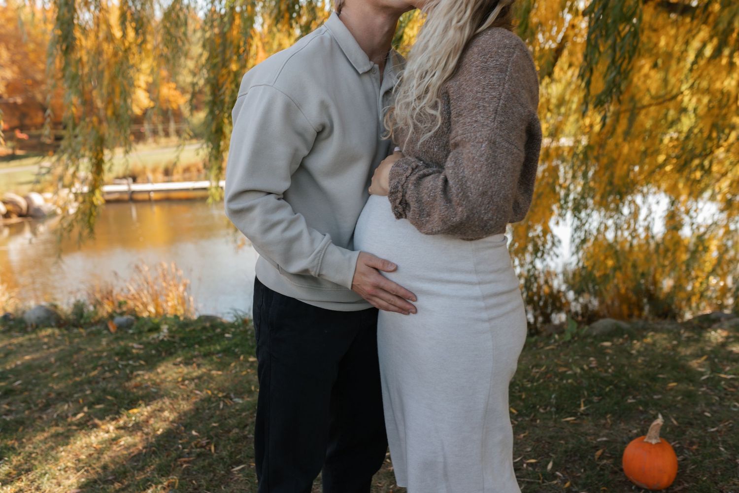Couple embracing outdoors in autumn, showing pregnant belly near a pond with fall foliage and a pumpkin nearby.