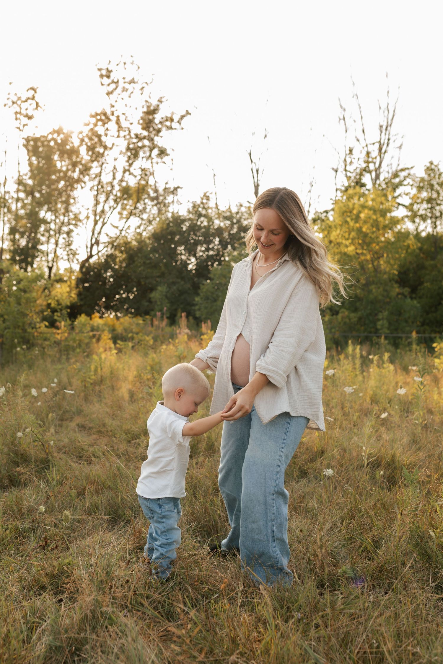 Pregnant woman in beige outfit smiling at toddler son in a sunlit golden meadow during sunset.