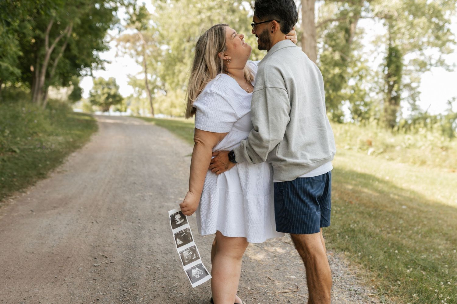Couple kissing on rural dirt road while woman holds ultrasound photos announcing pregnancy.