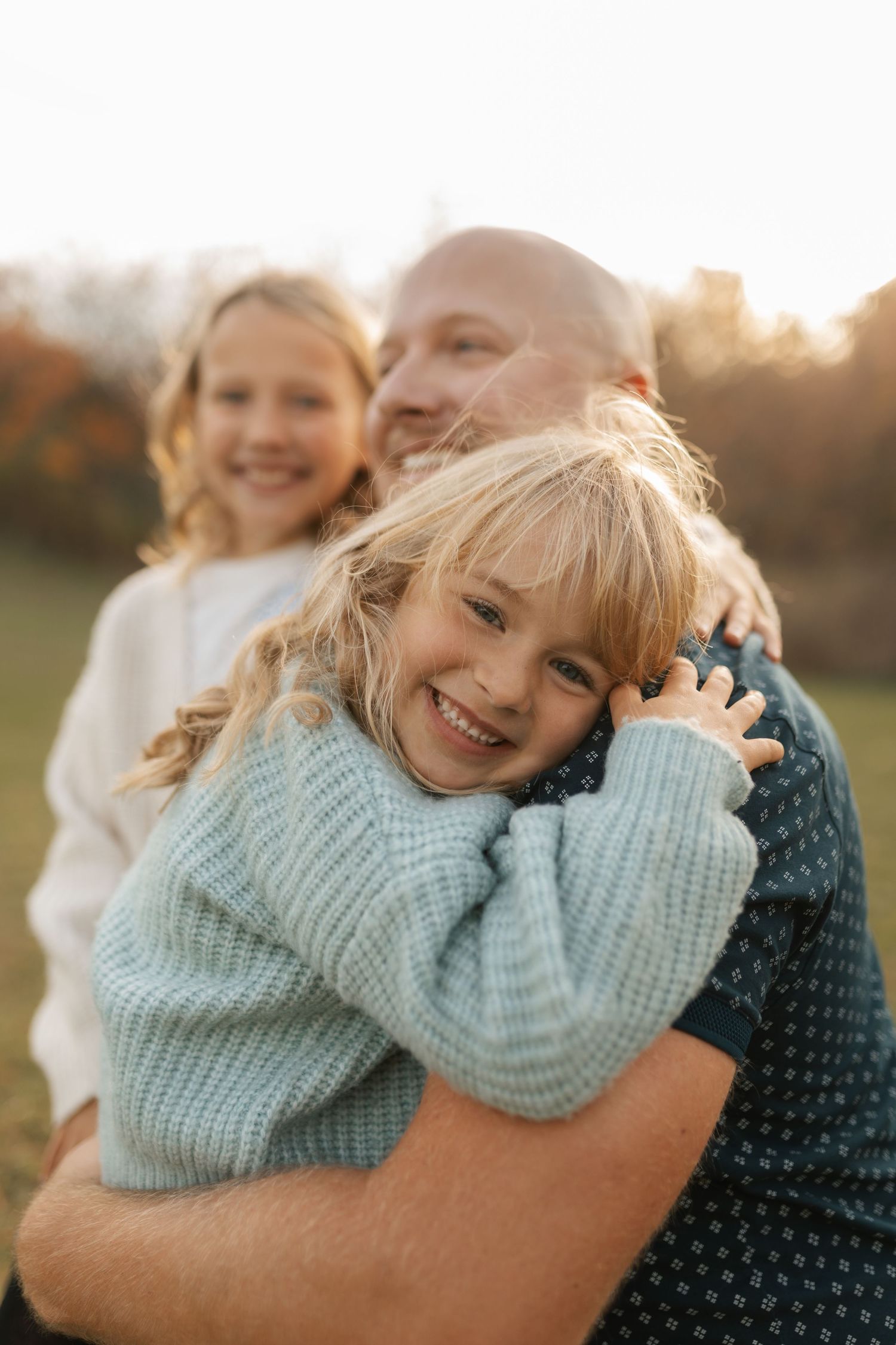 Young girl in teal sweater hugging her bald father outdoors in a sunny autumn field, smiling joyfully.