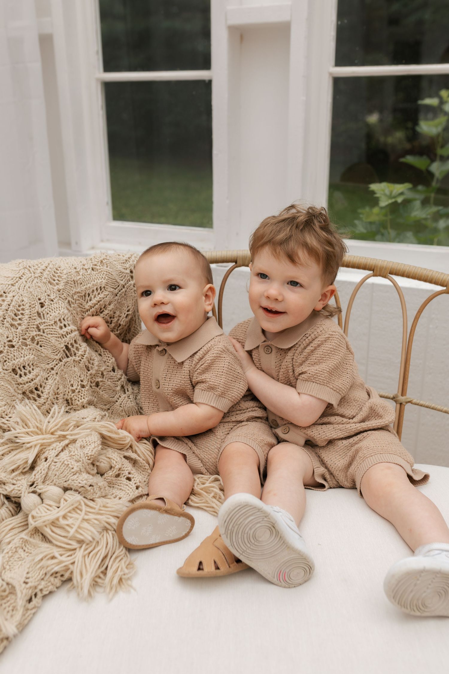 Two babies in matching beige rompers sitting together on a rattan bed with a knit blanket.