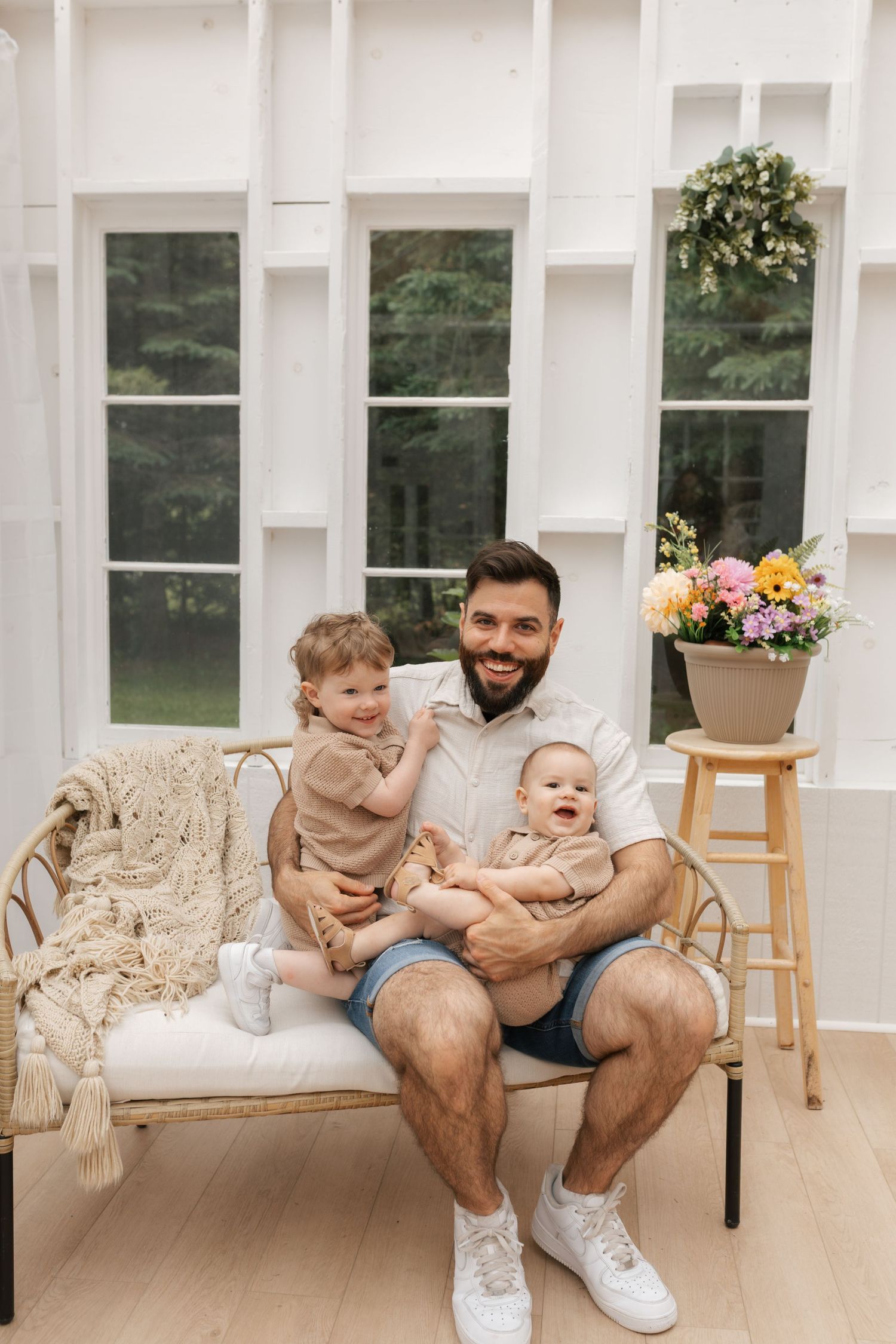 A smiling father sits on a wicker bench holding two toddlers in a bright white sunroom with flowers.