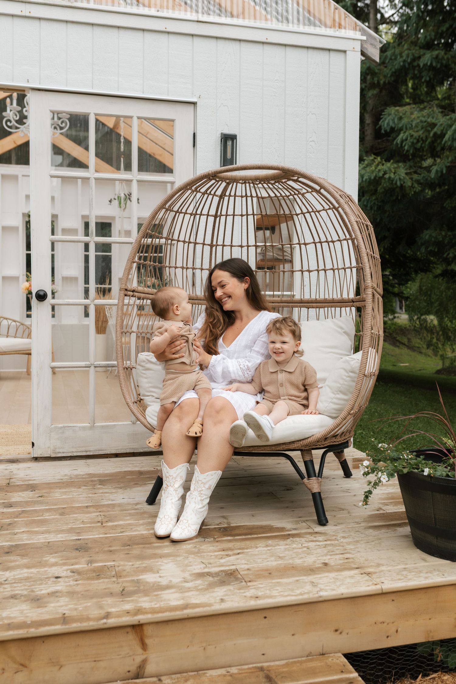 A woman in a white dress sits in a rattan egg chair on a wooden deck with two toddlers.