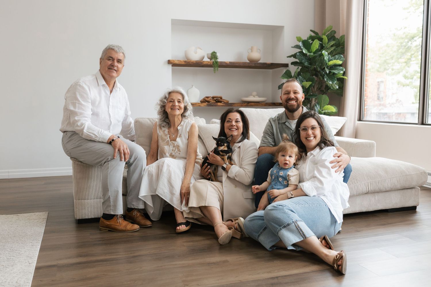 Multi-generational family portrait with grandparents, parents, baby, and dog on white sofa.