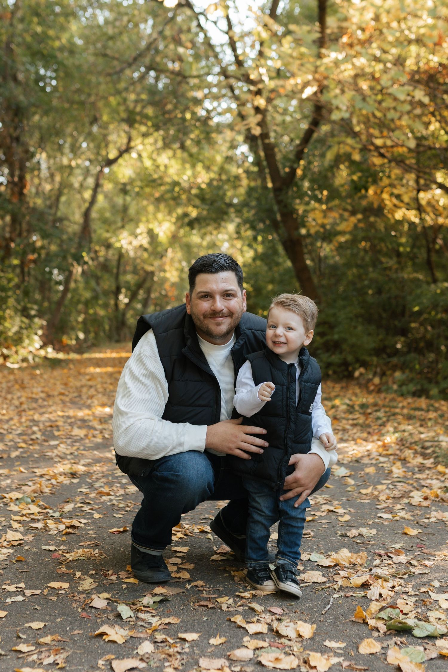 Father crouching beside toddler son on a leaf-covered path in an autumn forest, both smiling at camera.