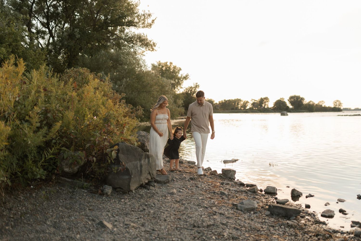 Pregnant couple walking along a rocky riverbank at golden hour, surrounded by lush green trees.