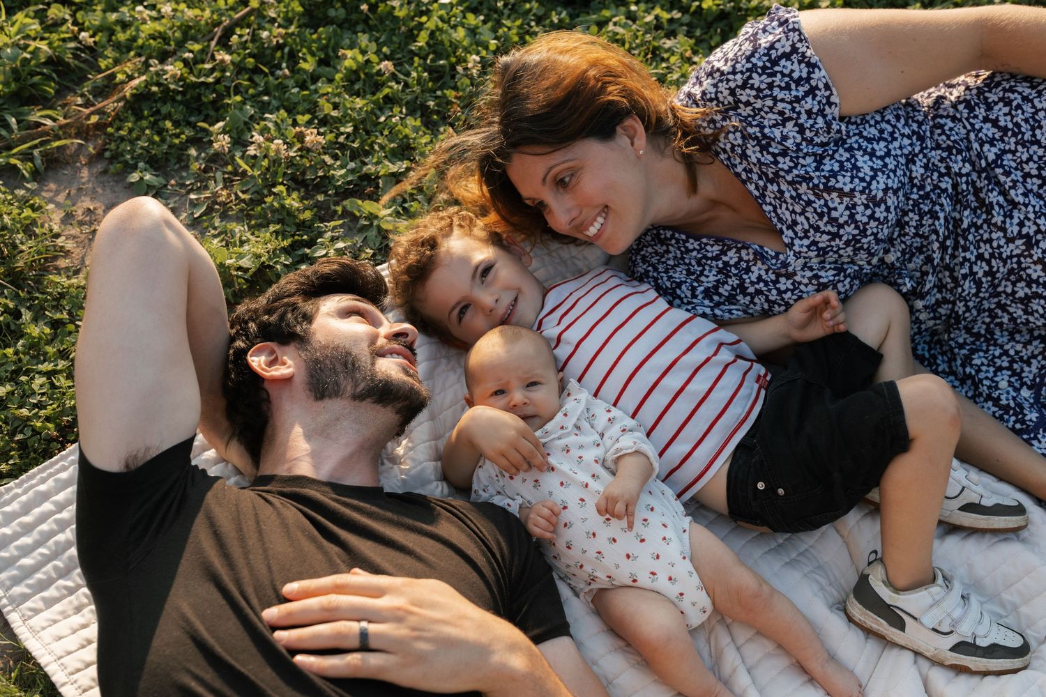 Young family of four relaxing on a blanket outdoors, parents and two children lying together smiling.