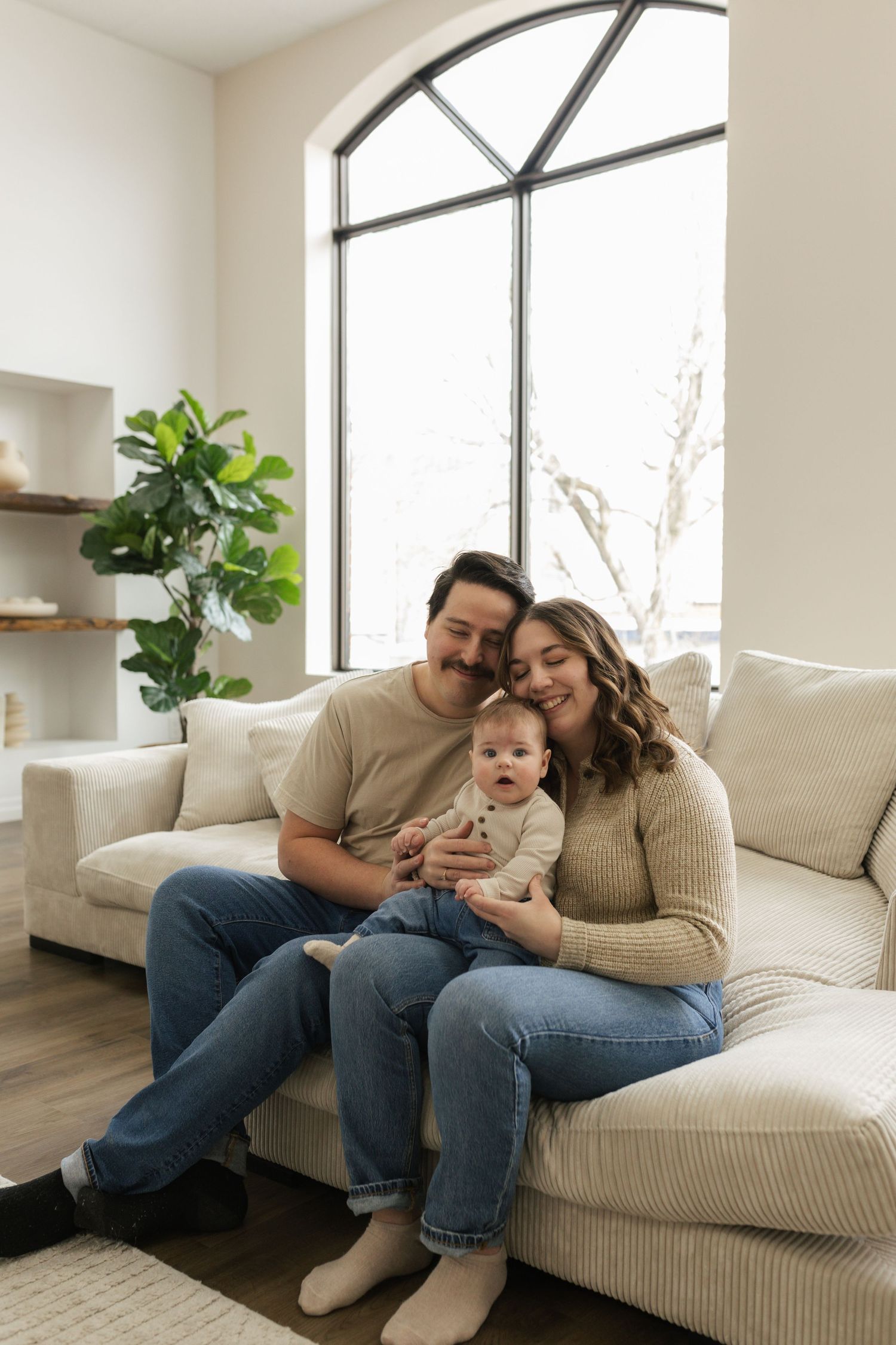 Happy young family with baby sitting on white couch in bright modern living room with large arched window.