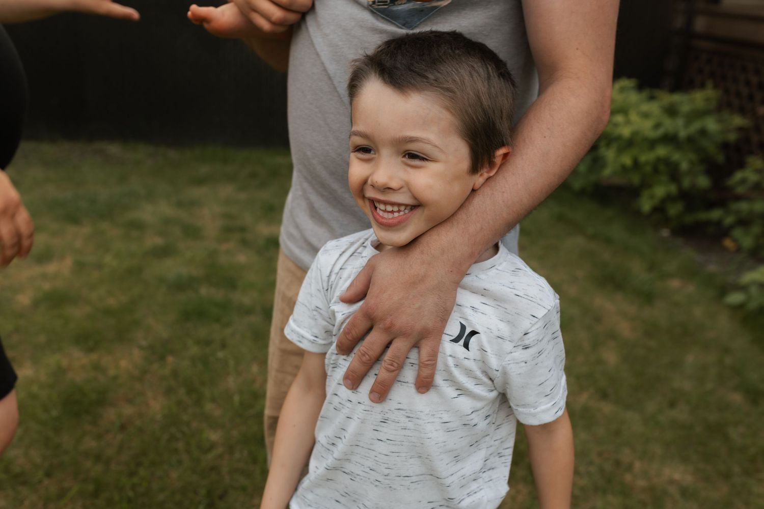 Smiling young boy in a white Hurley shirt laughing outdoors while an adult places a hand on his shoulder.