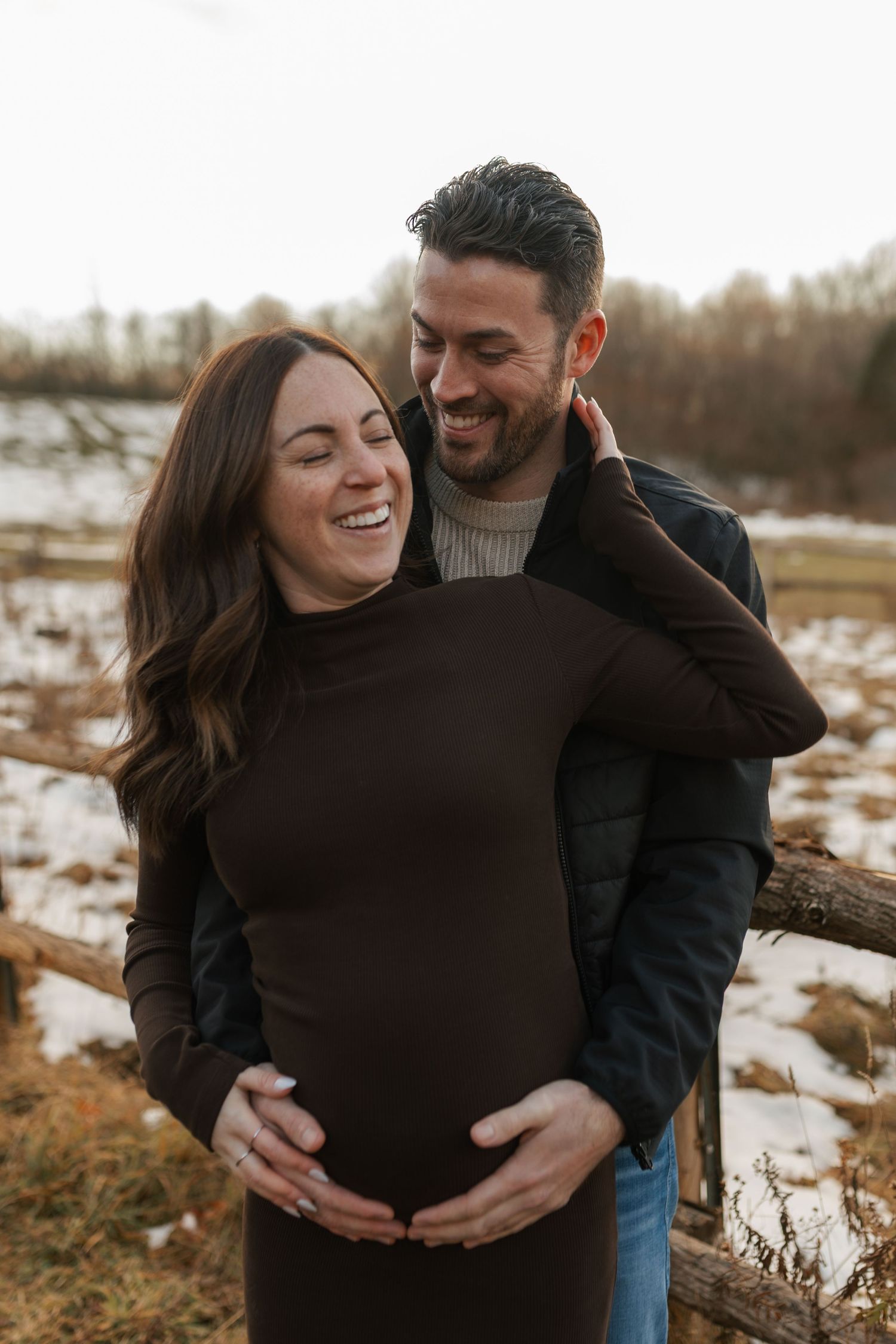 Happy pregnant couple embracing outdoors in winter, smiling together with snowy landscape in the background.