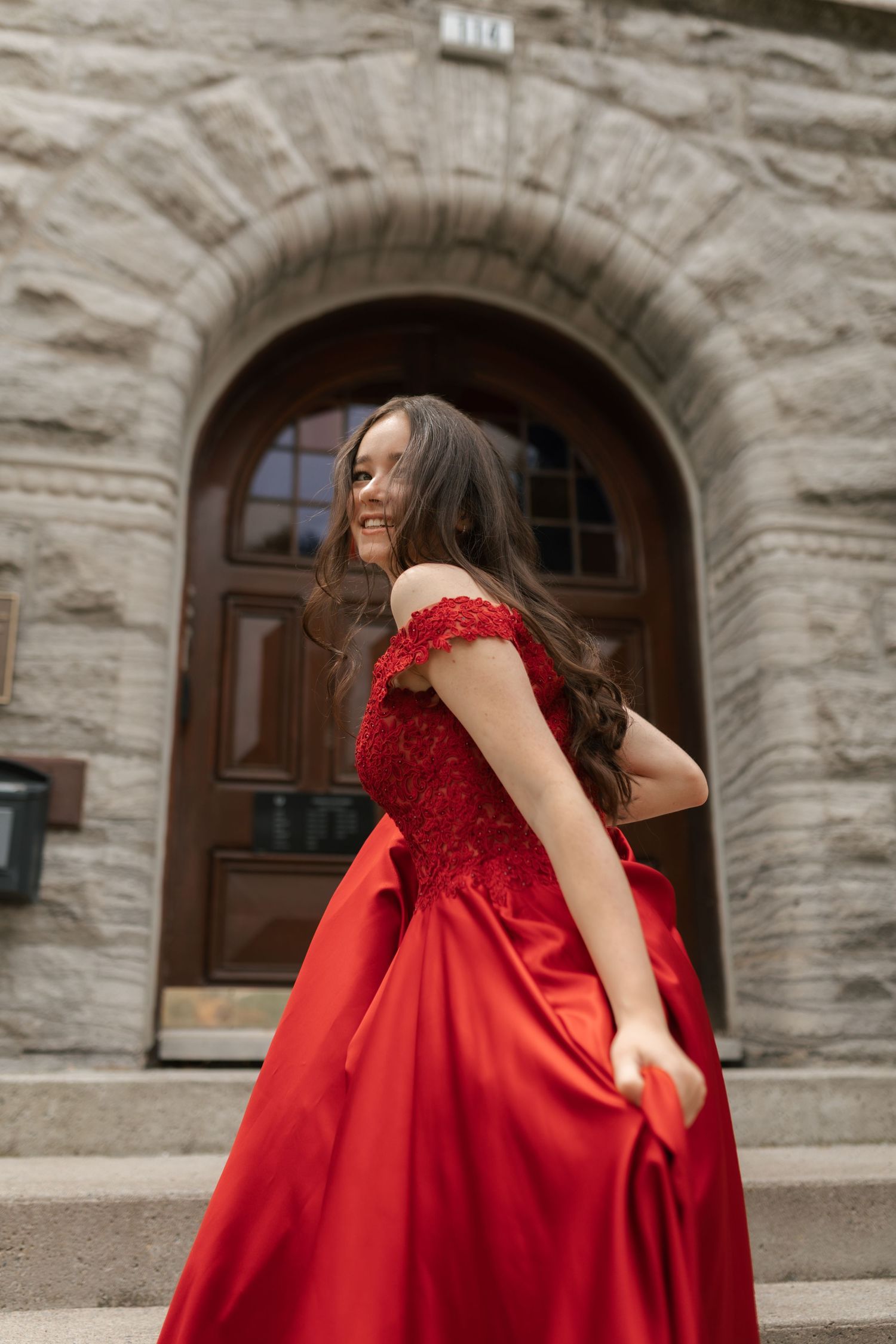 Young woman in elegant red off-shoulder ball gown twirling in front of a stone building with arched doorway.