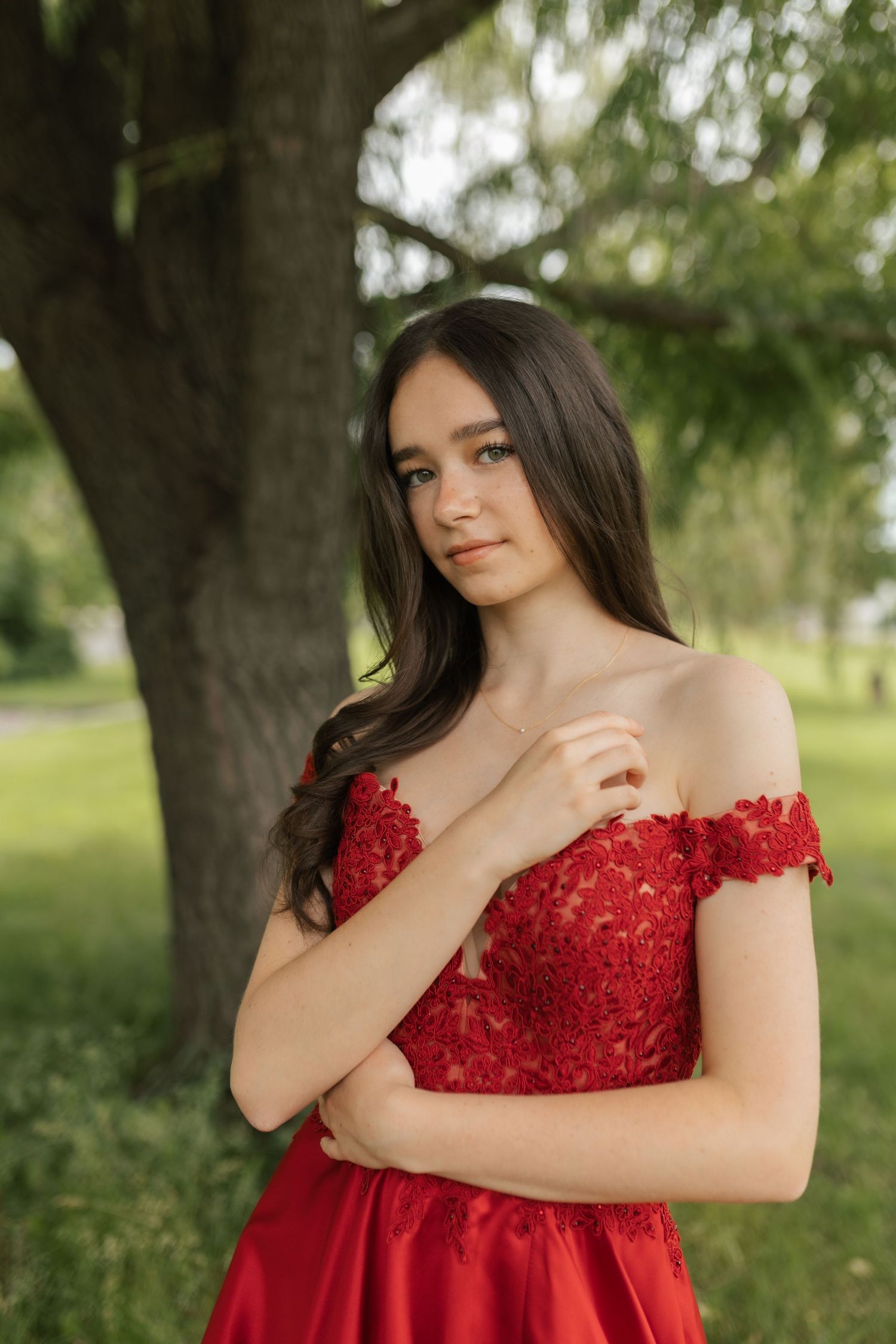 Young woman in red off-shoulder lace gown poses gracefully outdoors near a large tree in a lush green park.
